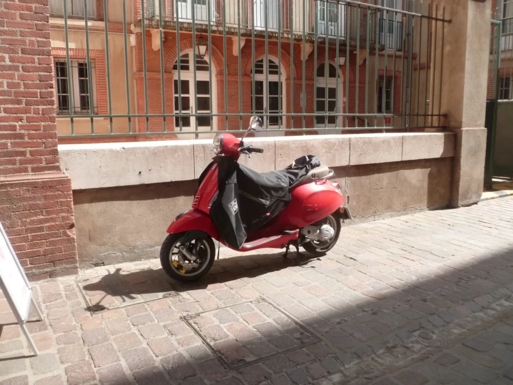 Un scooter rouge garé le long d’un immeuble en brique, rue pavée