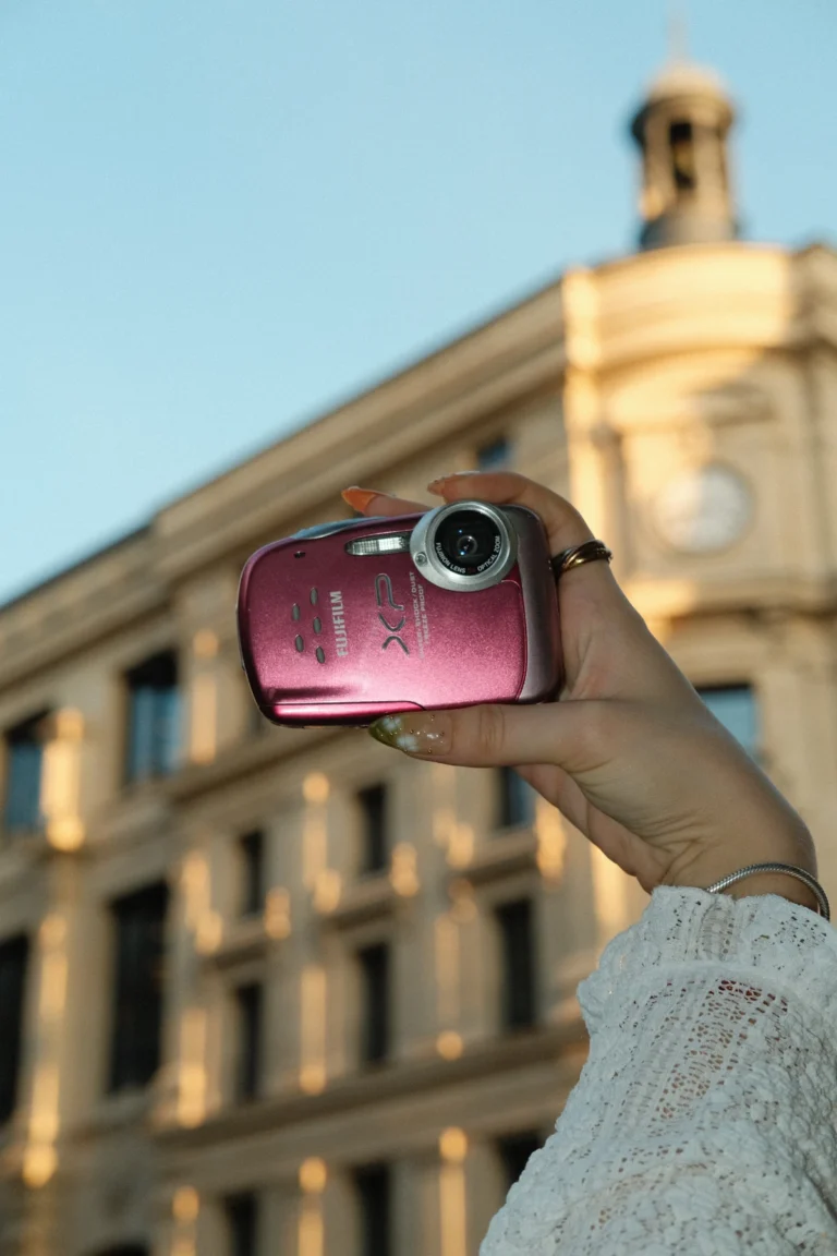 Le Fujifilm FinePix XP15 rose tenu à bout de bras devant un immeuble haussmannien, à Paris