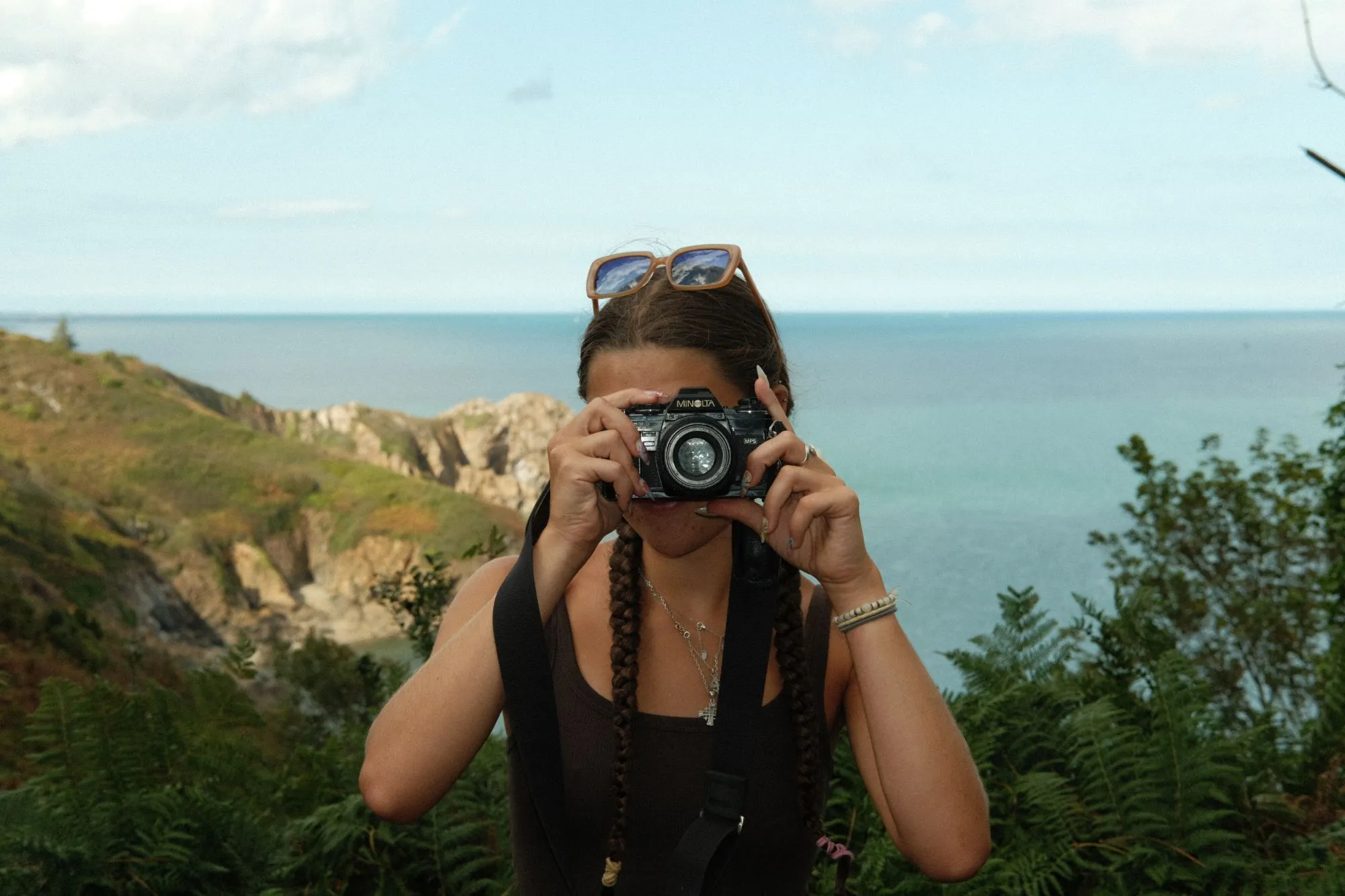 Un reflex argentique Minolta tenu à hauteur d’œil face à la mer, sur un sentier côtier bordé de fougères